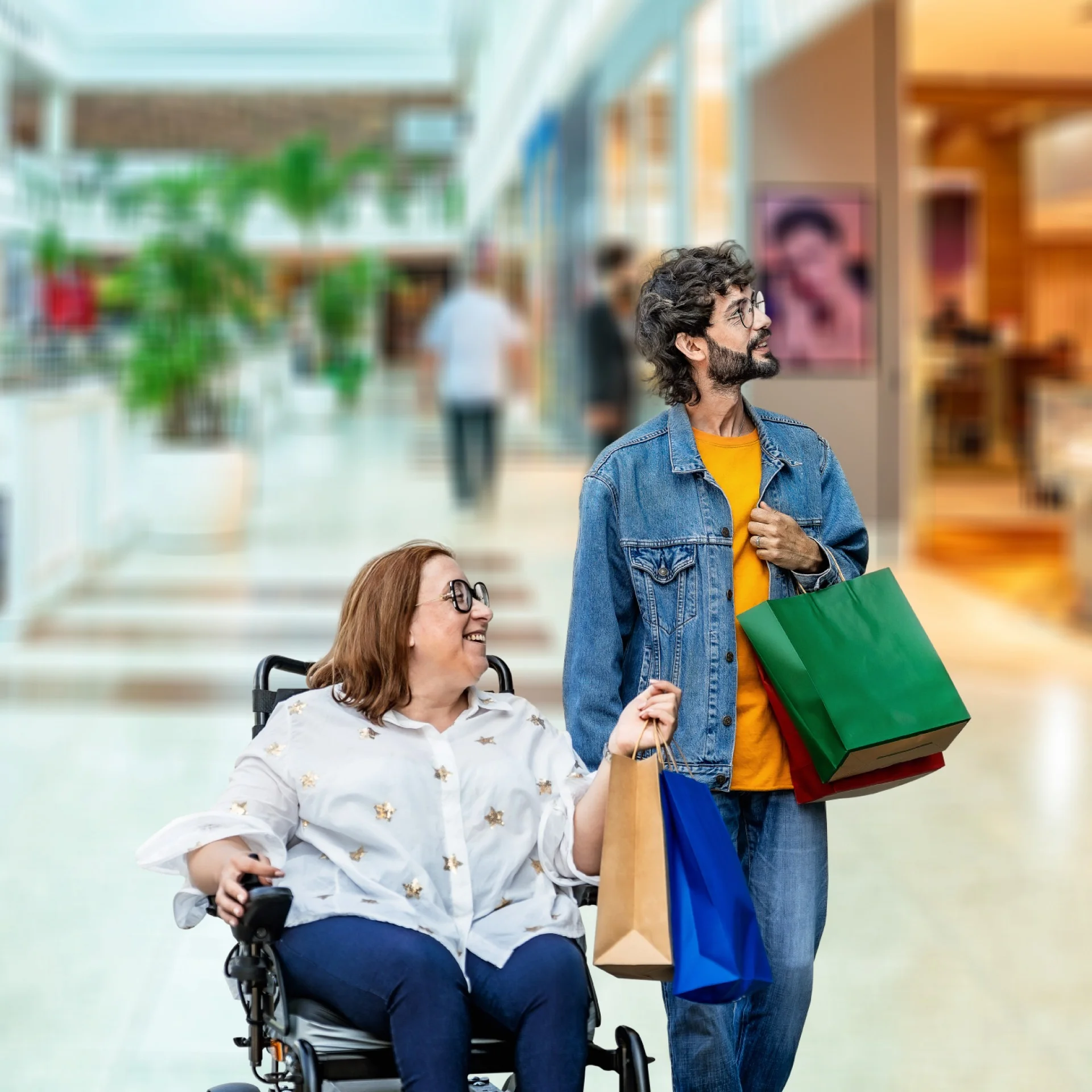 Duas pessoas passeando no Shopping sendo que uma delas está em uma cadeira de rodas apontando para uma loja. A outra está com uma sacola